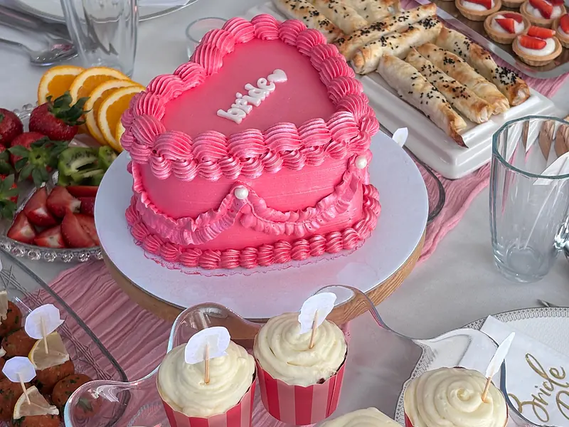 Mesa de postres para despedida de soltera con pastel de corazón rosa, cupcakes y fruta